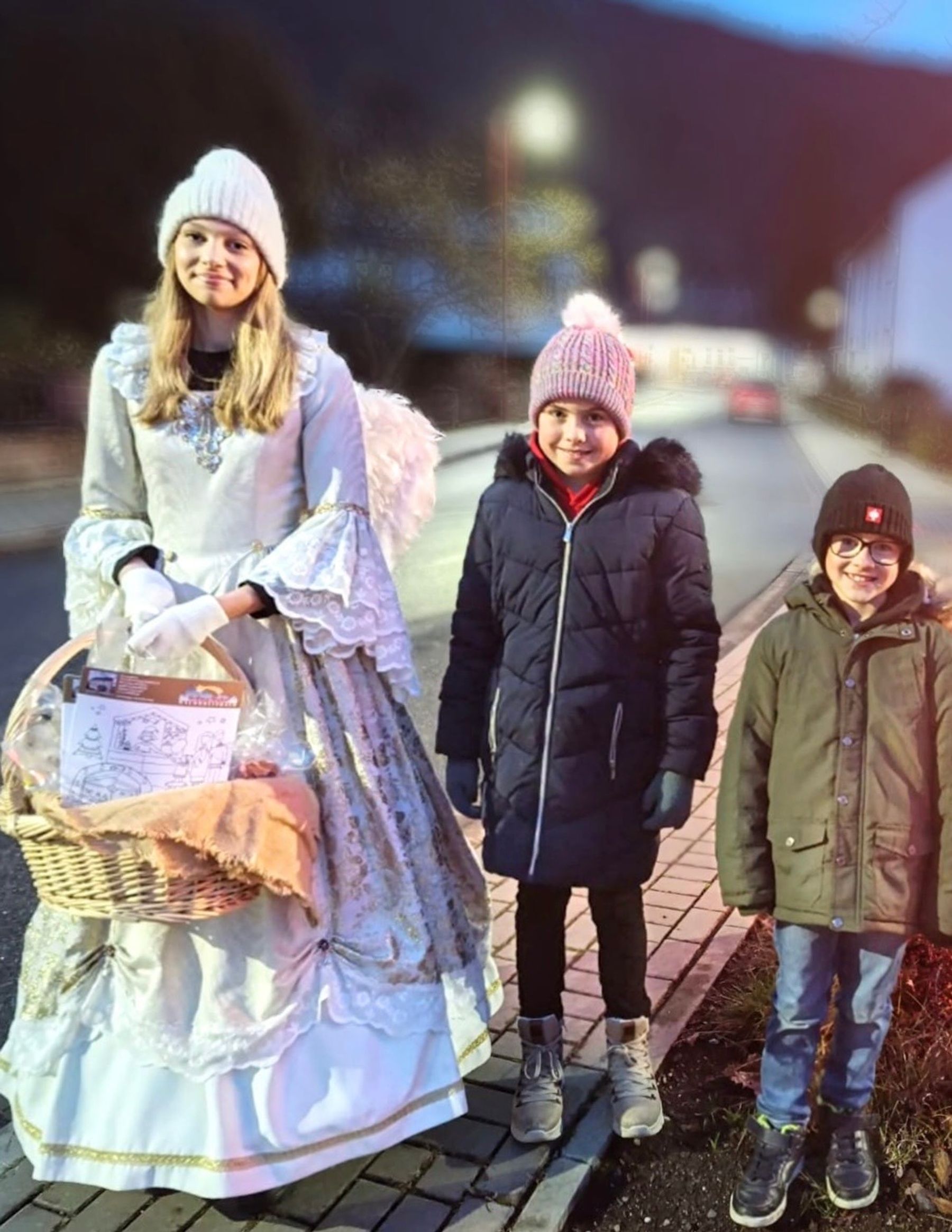 A young woman dressed as an angel with wings and a white costume holds a basket, standing on a sidewalk next to two smiling children in winter jackets and knit hats; it’s evening and the background shows a blurred street.